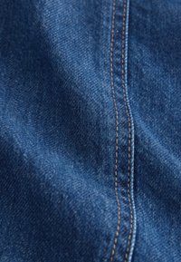 Tissu en denim bleu avec texture visible et une couture centrale double en fil beige, montrant de légers plis et ombres.