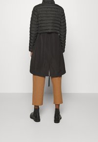 Chaqueta negra acolchada recortada sobre un vestido negro largo, combinada con pantalones marrones recortados y botines negros. Textura mate, diseño sencillo.