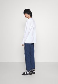 Camisa blanca de manga larga combinada con vaqueros azules holgados y sandalias deportivas en blanco y negro, de pie contra un fondo neutro.
