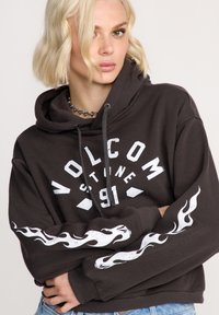 Hoodie gris foncé avec une graphique de flammes blanches sur les manches, grand logo de la marque sur le devant, capuche avec cordons de serrage, et coupe décontractée.