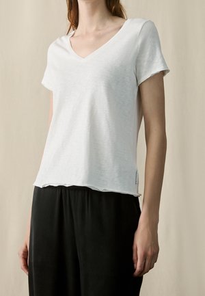 Kobieta ubrana w gładką białą koszulkę z krótkim rękawem i dekoltem w serek, w połączeniu z luźnymi czarnymi spodniami, stojąca na neutralnym tle.