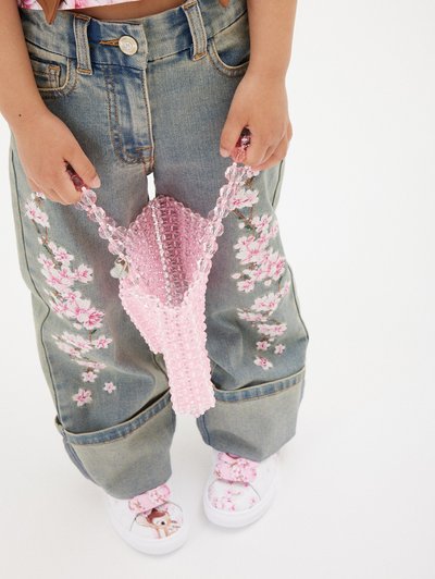 Borsa rosa con perline e manico ad arco, tenuta da un bambino che indossa jeans di denim ricamati con motivi floreali e scarpe con motivi floreali.
