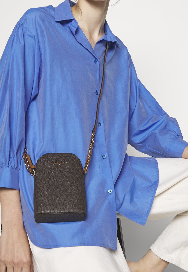 Brązowa torebka crossbody o wzorzystym wykończeniu, z łańcuszkowym paskiem i okrągłym dnem, zestawiona z niebieską koszulą z guzikami i białymi spodniami.