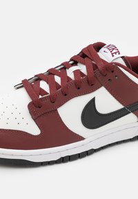 Nike sneakers med en vit läderbas, maroon syntetiska detaljer, svart swoosh och strukturerade maroon snören. Gummisula med greppmönster.