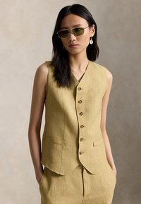 Ασιάτισσα γυναίκα που φοράει αμάνικο γιλέκο με κουμπιά σε ελαιόχρωμο πράσινο, ταιριαστά παντελόνια, γυαλιά ηλίου με χρυσή κορνίζα και λευκά σκουλαρίκια που κρέμονται, με τα χέρια στις τσέπες.