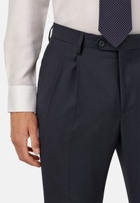 Navy taisītās bikses ar bodeņiem, izgatavotas no teksturēta auduma. Ar pogu aizdari un šauru, strukturētu piegriezumu. Redzama baltu krekla piedurkne.