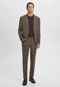 Bruin op maat gemaakt pak met een enkel knoopjack en rechte broek, gecombineerd met een donkere top. Het heeft een gestructureerde stof en een slim fit.