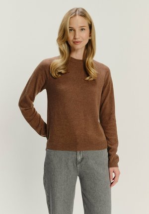 Blondynka z luźnymi falami nosi brązowy sweter z długim rękawem i szare dżinsy, stojąc z jedną ręką za plecami.