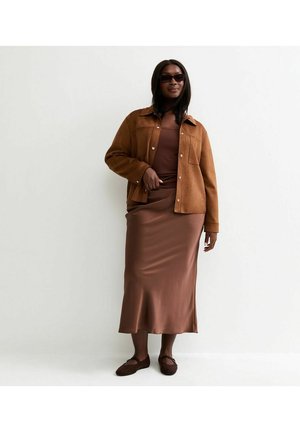 Femme portant une robe midi marron, une veste boutonnée beige, des lunettes de soleil foncées et des ballerines marron, debout devant un fond blanc.
