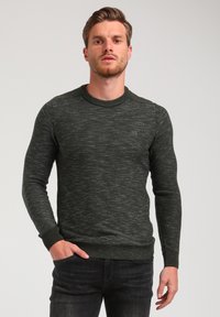 Pull en tricot vert foncé avec un motif texturé, col ras du cou et poignets côtelés. Détail logo discret sur la poitrine gauche.