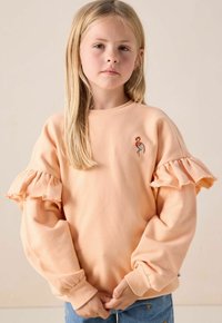 Blond meisje in een perzikkleurige sweatshirt met gerimpelde mouwen en een kleine flamingo-borduring, staand met haar handen samengevouwen.
