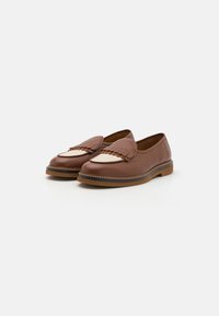 Zapatos marrones de piel tipo mocasín que presentan un detalle de flecos en forma de ondas y una sección beige contrastante en la parte superior. Puntera redondeada y suela plana.
