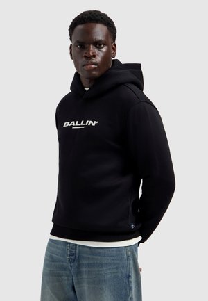 Jonge man draagt een zwarte "BALLIN'" hoodie en een blauwe spijkerbroek, staand tegen een effen lichtgrijze achtergrond, kijkt recht vooruit.