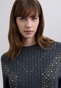 Maglione grigio a coste con borchie metalliche dorate e argentate disposte in un motivo decorativo lungo la parte anteriore e le spalle.