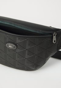 Sac banane en simili cuir noir avec motif géométrique en relief, fermeture zippée et logo Lacoste. Bretelle ajustable pour un port sécurisé.