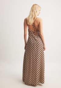Maxi dress with a brown and beige striped design. Features a low back with crisscross straps and a tied detail. Smooth fabric texture.
