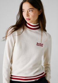 Maglione crema con colletto e orlo a coste a strisce rosse e bianche; presenta il logo ricamato "Red Bull RACING" sul petto, texture morbida.