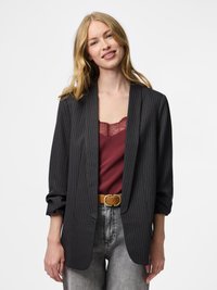 Junge Frau mit langem blondem Haar, die einen schwarzen Nadelstreifen-Blazer, ein burgunderrotes, mit Spitze verziertes Oberteil, einen Hellbraunen Gürtel und hellgraue Jeans trägt und lächelt.