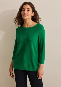 Grön stickad tröja med rund halsringning, trekvartsärmar och en centralseamdetalj. Mjuk textur, avslappnad passform. Matchad med mörka jeans.