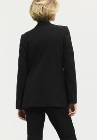 Sort skræddersyet blazer med spidse klapper, strukturerede skuldre og knapdetaljer, kombineret med matchende skræddersyede bukser. Glat stoftekstur.