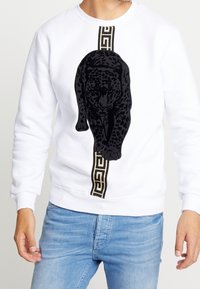 Hombre vistiendo un sudadera blanca con un gráfico de pantera negra, caminando por una franja vertical de patrones en oro y negro, combinada con unos jeans de color azul claro.