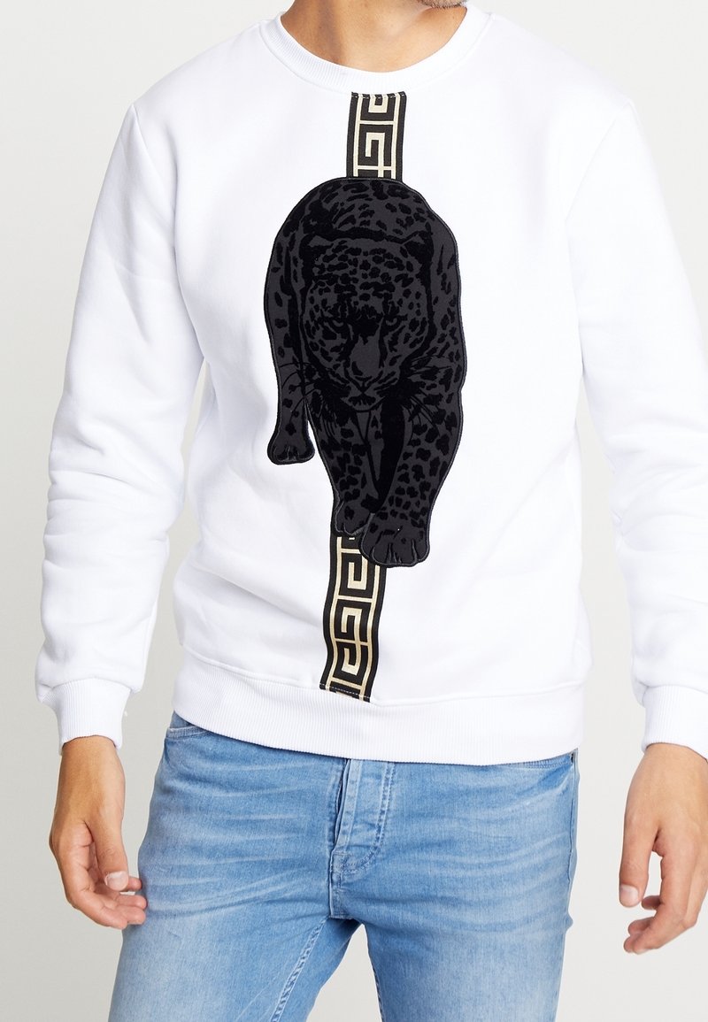 Hombre vistiendo un sudadera blanca con un gráfico de pantera negra, caminando por una franja vertical de patrones en oro y negro, combinada con unos jeans de color azul claro.