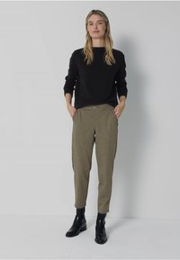Zwarte lange mouwen trui in combinatie met olijfgroene slim fit broek. Broek heeft zijzakken. Model draagt zwarte enkellaarsjes. Effen achtergrond.