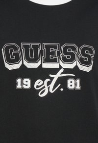 Černé tričko s nápisem "GUESS" v tučném, bílém, obrysovém písmu a s "est. 1981" ve stříbrných akcentů na přední straně.