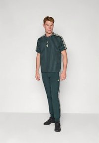 Vyras stovintis prieš baltą foną, dėvintis tamsiai žalią trumpomis rankovėmis marškinius su dryžiais ir atitinkamas sportines kelnes su baltomis juostelėmis bei juodus batus.