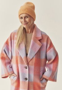 Villa mantel pastelses ruudumustris, millel on suured kraed, kaks esimest taskut ja puidust nööbid. Kombineeritud tekstuurse oranži kudumiga mütsiga.