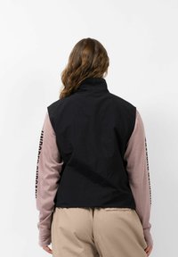 Schwarze Stoffaußenschicht mit hohem Kragen, kombiniert mit hellrosa, gerippten Ärmeln mit Aufdruck. Beigefarbene Hose vervollständigt das Outfit.
