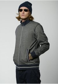 Giacca zip-up a scacchi grigia con colletto alto e dettagli in blu navy. Presenta polsini e vita a coste, con due tasche laterali.