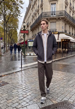 Jeune homme marchant sur une rue pavée humide près de l'entrée du métro et d'un café dans une ville européenne avec des arbres d'automne et des bâtiments historiques.