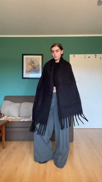 Châle noir à franges fait de tissu épais, drapé sur les épaules, avec des extrémités lâches qui pendent. Porté avec un pantalon large gris.