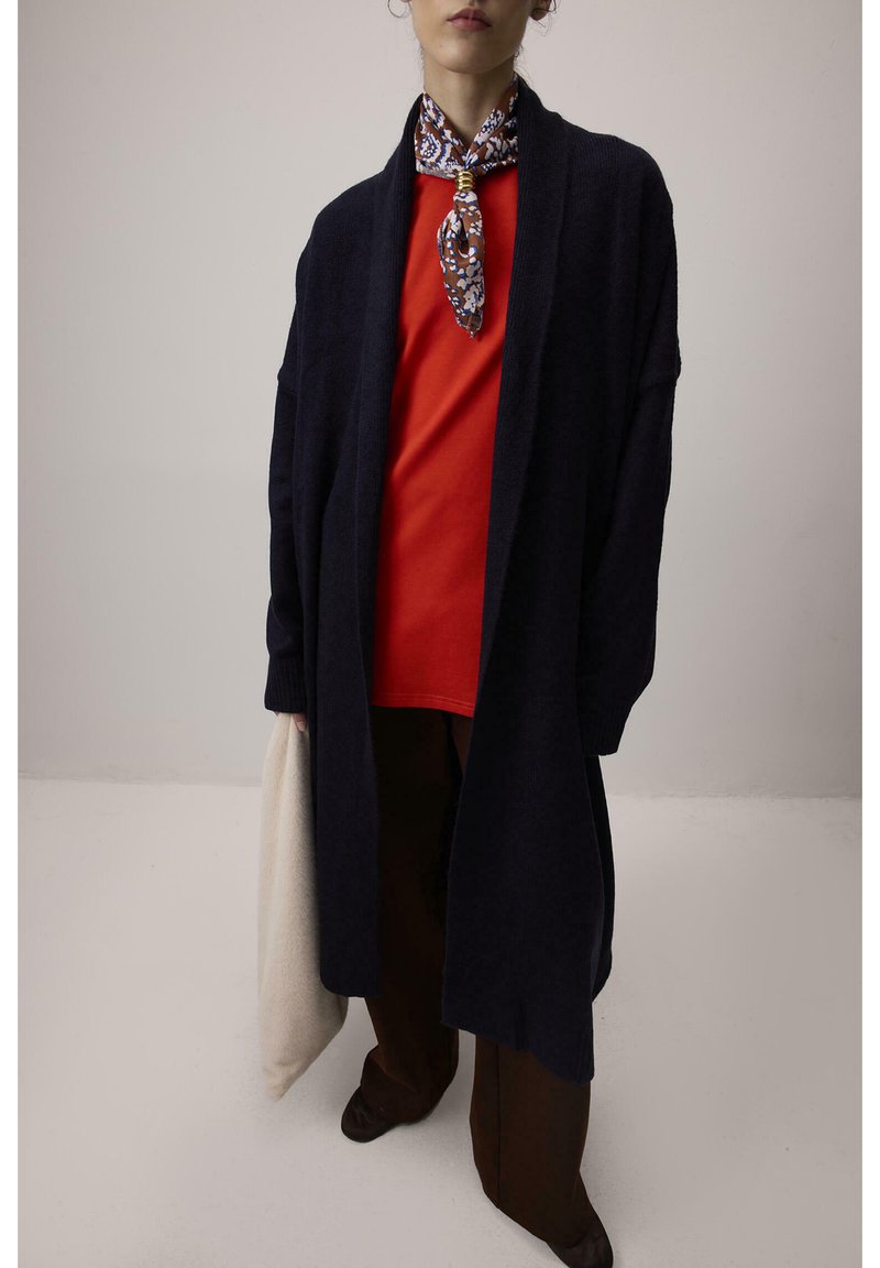 Άτομο φορά μακρύ σκούρο ζακέτα, φωτεινό κόκκινο πουκάμισο, σκούρο παντελόνι, φουλάρι με σχέδια στο λαιμό, κουβαλώντας ανοιχτόχρωμη τσάντα μπροστά σε απλό φόντο.