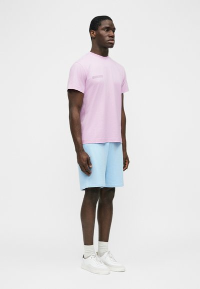 Άνδρας που φοράει ανοιχτό μωβ μπλουζάκι, ανοιχτό μπλε σορτς, λευκές κάλτσες και λευκά αθλητικά παπούτσια, σε απλό λευκό φόντο.