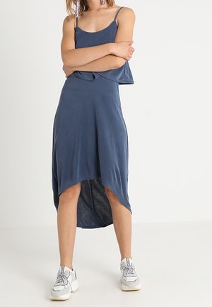 Woman standing with arms crossed wearing a sleeveless blue asymmetrical midi dress and white sneakers on a plain background.