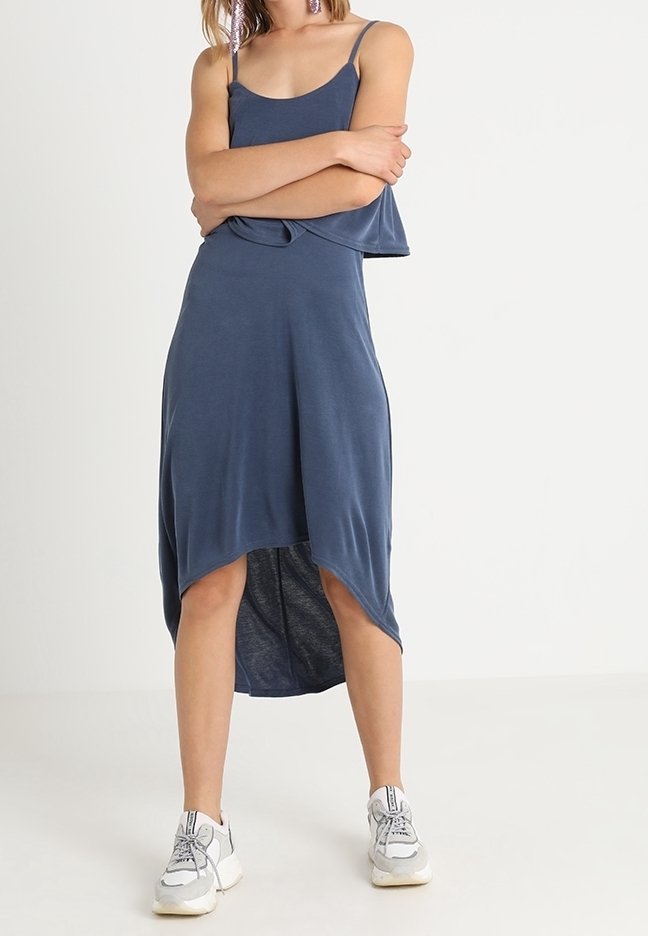 Femme debout, les bras croisés, portant une robe midi asymétrique bleu sans manches et des baskets blanches sur un fond uni.