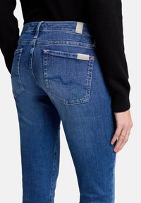 Dark blue denim jeans featuring a mid-rise design, back pockets with stitching details, and a smooth texture. Silver hardware accents are visible.