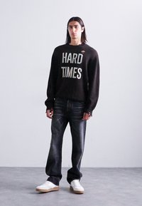 Junger Mann mit langem Haar, der einen schwarzen Pullover mit dem Schriftzug "HARD TIMES", dunkle Jeans und weiße Sneakers trägt und vor einer schlichten weißen Wand steht.