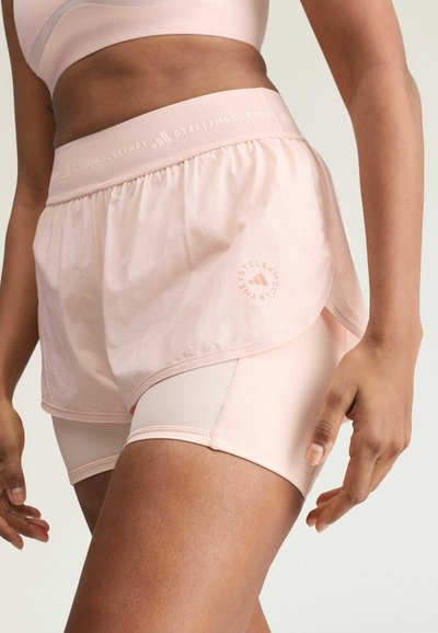 Frau, die hellrosa geschichtete sportliche Shorts und einen passenden Sport-BH trägt, gezeigt vom Bauch bis zu den oberen Oberschenkeln vor einem einfarbigen Hintergrund.