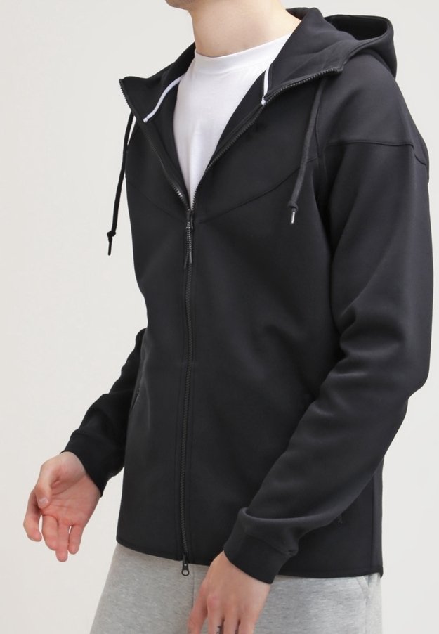 Person som bär en svart zip-up hoodie med dragsko över en vit skjorta och ljusgrå byxor, stående mot en enkel bakgrund.