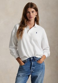 Junge Frau mit langen Haaren, die ein weißes Langarm-Poloshirt und blaue Jeans trägt, Hände in den Taschen, neutraler Hintergrund.