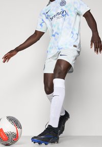 Mies jalkapalloilija valkoisessa ja sinisessä pelipaidassa hallitsee mustaa ja valkoista Nike-palloa, jossa on punainen swoosh, yksinkertaisella taustalla.