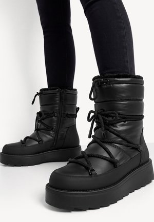 Bottes d'hiver noires matelassées avec semelles épaisses, lacets en corde enroulés, fermetures éclair latérales, portées avec un pantalon noir ajusté.