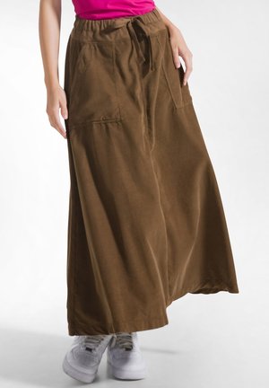 Persoon draagt een lange bruine rok met voorzakken, witte sneakers en een roze top zichtbaar bij de taille.