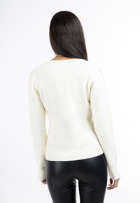 Maglione di colore crema con maniche lunghe, polsini e orlo a costine. Sul retro presenta un scollo a barchetta moderato. Abbinato a pantaloni neri aderenti.