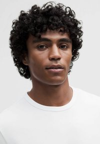 Portrét osoby s kudrnatými černými vlasy, která má na sobě jednoduché bílé tričko, na pozadí světle šedé barvy. Čistá pleť a neutrální výraz.