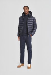Giacca a vento imbottita blu navy con cappuccio, design trapuntato, due tasche laterali con zip; indossata sopra un maglione blu navy, abbinata a pantaloni blu navy e sneakers marroni.