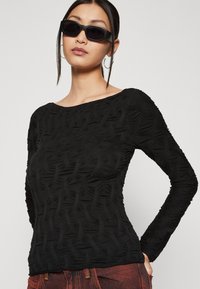 Zwarte longsleeve top met een gestructureerd patroon, ronde halslijn en aansluitende pasvorm. Gecombineerd met zwarte zonnebril. Achtergrond is effen wit.
